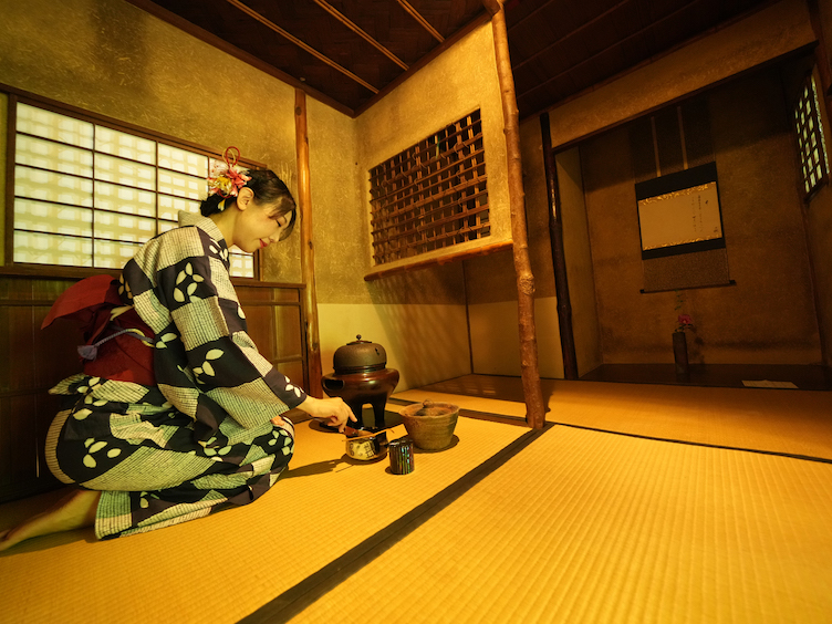 Eine Besucherin eines traditionellen japanischen Hauses kniet im Yukata gekleidet in einem Tatami-Zimmer und führt eine Teezeremonie durch.