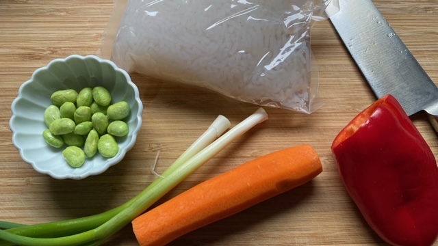 Konjak-Nudeln im Plastikbeutel, Karotte, Schälchen mit Edamame, Lauchzwiebel und rote Paprika auf Schneidebrett mit Messer