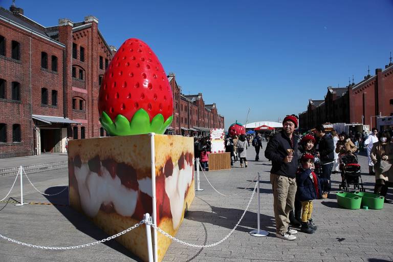 Riesenstück Erdbeertorte aus Plastik vor roten Backsteinwarenhäusern mit Menschen im Hintergrund
