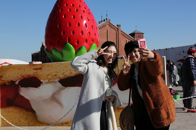 Zwei junge Frauen schießen Selfies vor einem Riesen-Stück Erdbeertorte aus Plastik