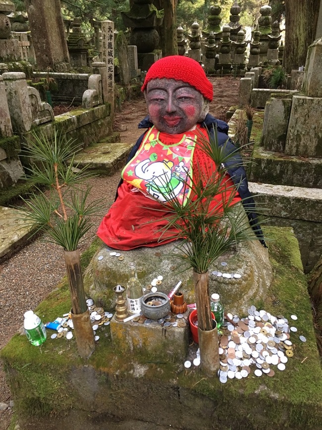 Jizo-Statur mit roter Kleidung und von Opfergaben umgeben.