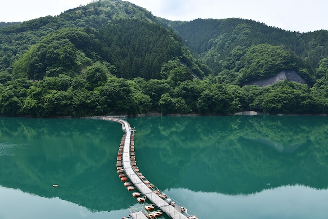 Mugiyama-Schwimmbrücke auf dem grünlichen Okutama-See, auf dem sich die Berge im Hintergrund spiegeln