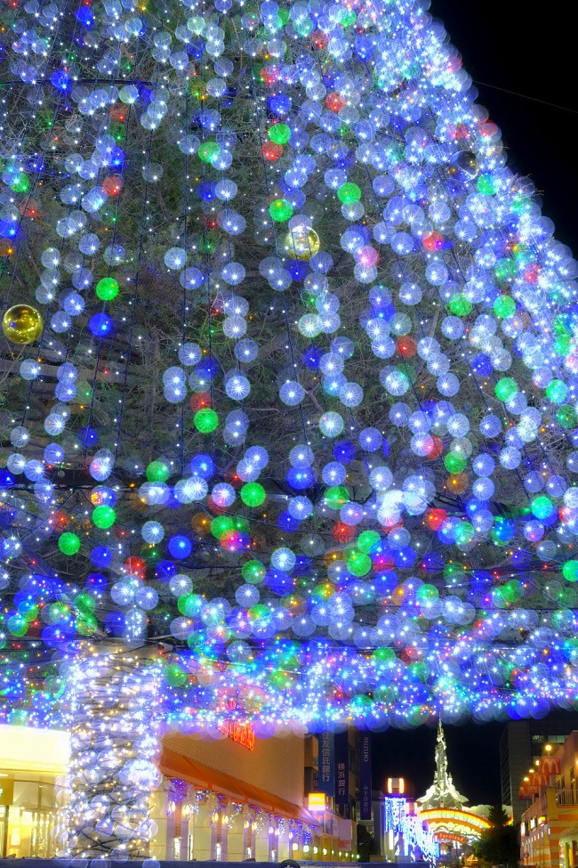 Nahaufnahme des beleuchteten Weihnachtsbaumes, im Hintegrund der beleuchtetete Eingang des Themenparks Sanrio Purioland