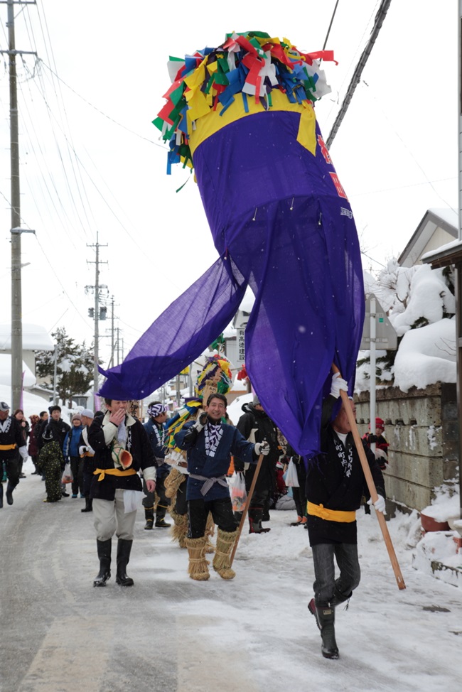 Eine Gruppe in festlicher Kleidung unterwegs in einer verschneiten Straße, vorneweg ein Manner, der das in lila Tuch und mit bunten Papiergirlanden geschmückte "bonden" trägt.