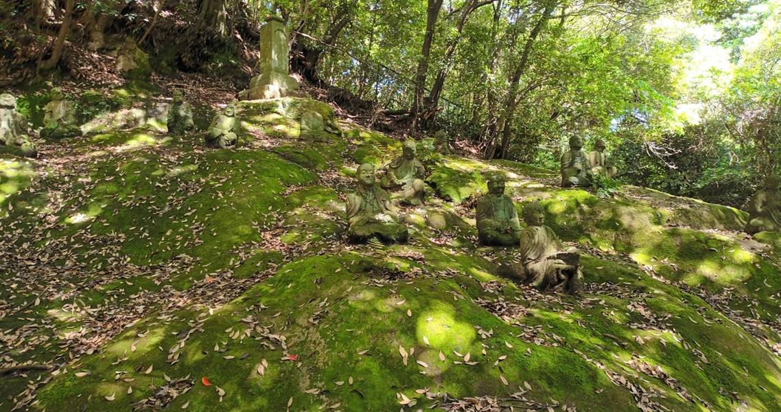 Buddha-Figuren auf moosbewachsenem Grund vor der Reigandō-Höhle