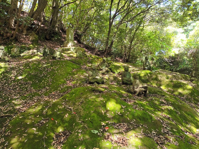 Buddha-Figuren auf moosbewachsenem Grund vor der Reigandō-Höhle