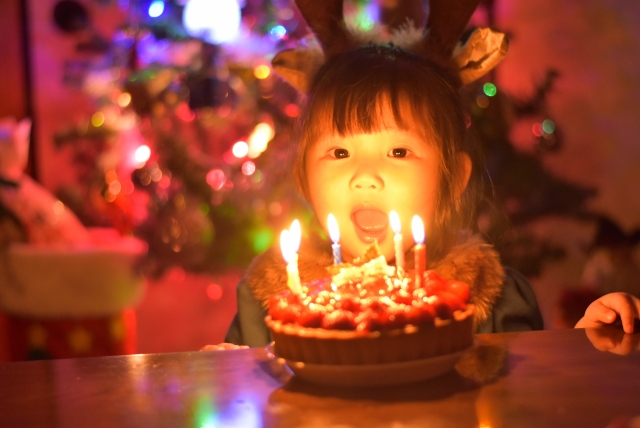 Kind bläst vor dem Weihnachtsbaum einen Kuchen mit Kerzen aus