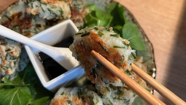 Daikon-Mochi wird mit Holzstäbchen über einen Teller mit Sauce, Salat und weiteren Mochi gehalten