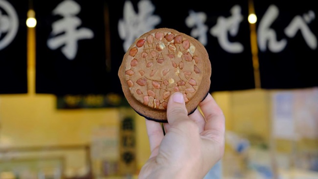 Hand hält einen Senbei vor einem Geschäft in die Höhe